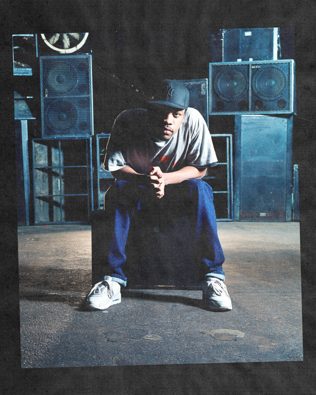 Wiley grime artist seated pose wearing air max 90 white staring at lens late in front of speaker wall in the 2000s